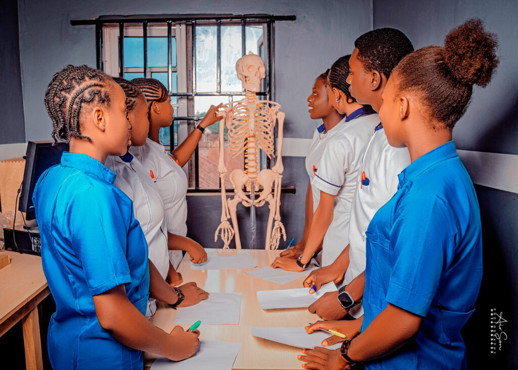 Anatomy Class in Session – Victory College Health Students in Practical Learning Group of Victory College health science students in uniforms gathered around a table with notebooks, engaging in an anatomy class with a human skeleton model
