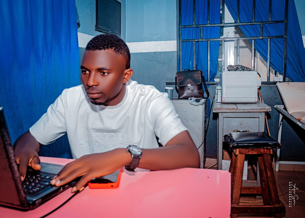 Registrar at Work – Victory College Administrative Office Male registrar working on a laptop at the Victory College administrative office, seated at a pink desk with office equipment in the background.